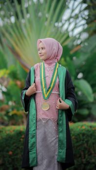 Young woman in graduation attire outdoors with foliage background, symbolizing achievement. - Fotografer Sidoarjo