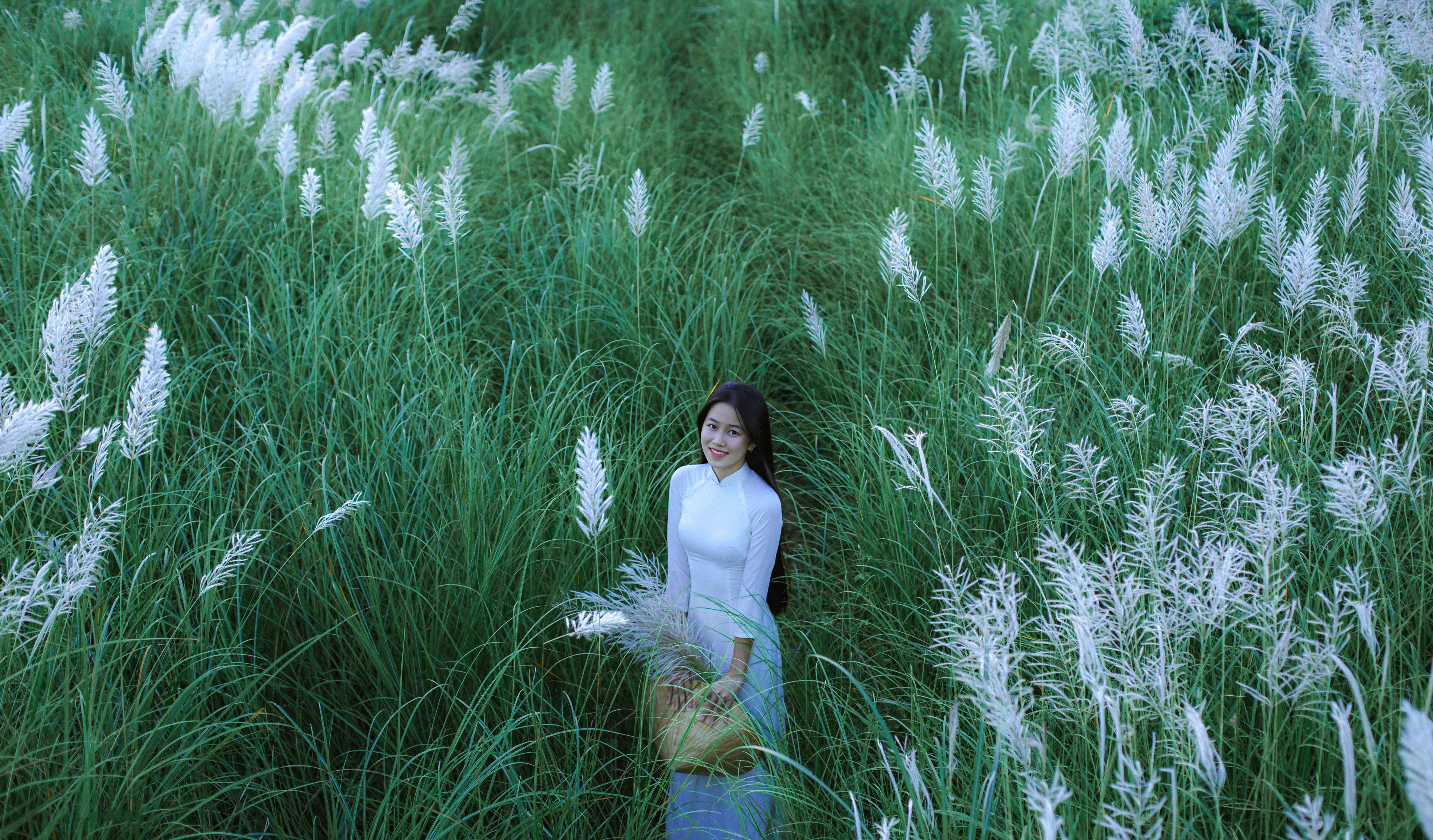 A woman in traditional Vietnamese dress standing in a lush green field with tall white flowers.