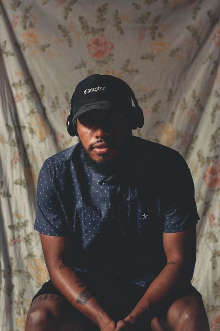 A stylish man with a beard, wearing headphones and a cap, poses seriously indoors.