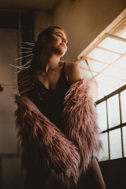 Stylish woman in a fur coat posing by a window with dramatic lighting.