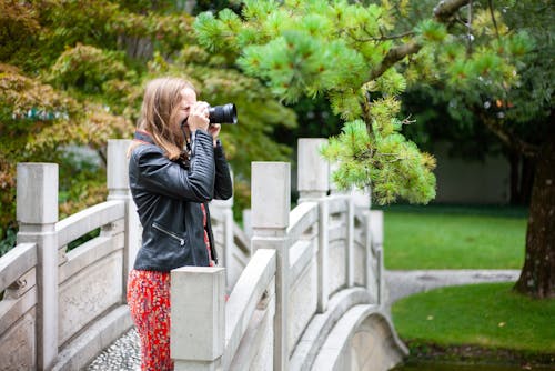 Free Woman Wearing Black Zip-up Jacket Holding Camera While Standing on White Bridge Stock Photo