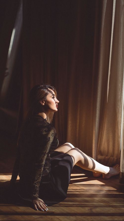 Free A young woman sits thoughtfully by a sunlit window with warm tones highlighting her contemplative mood. Stock Photo