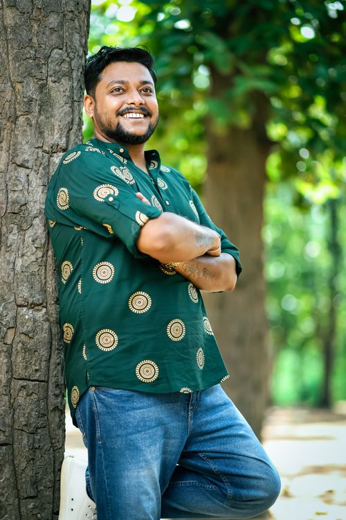 Free Portrait of a smiling man in a casual shirt leaning against a tree outdoors. Stock Photo