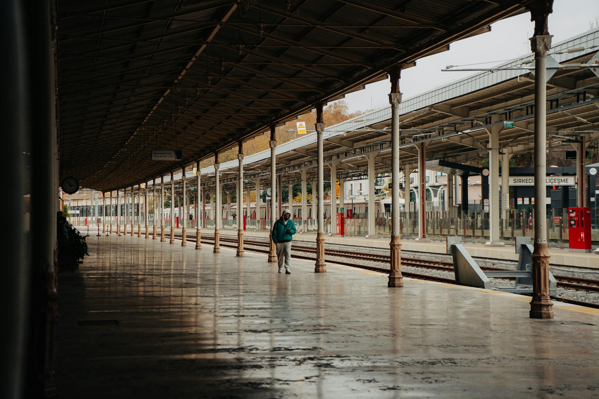 A lone person stands on an empty urban train station platform under cloudy skies.