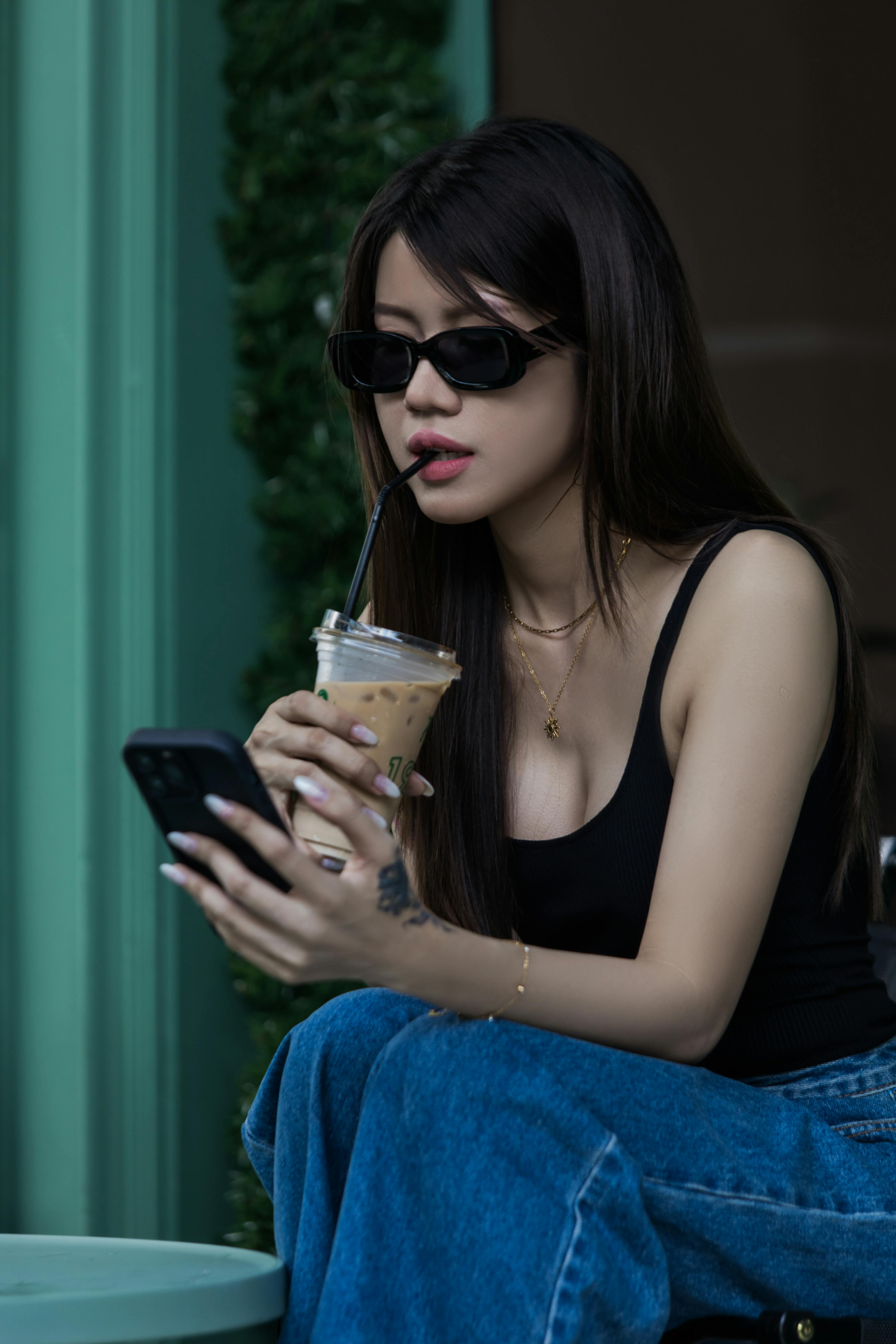 Miễn phí Người phụ nữ thời trang đeo kính râm đang nhâm nhi cà phê đá trong khi sử dụng điện thoại thông minh. Ảnh lưu trữ