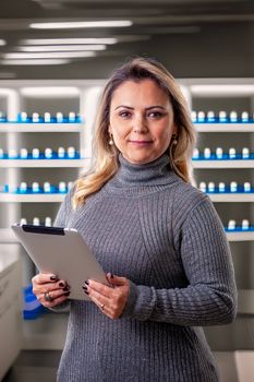Confident woman in a laboratory setting holding a tablet, surrounded by shelves with bottles.