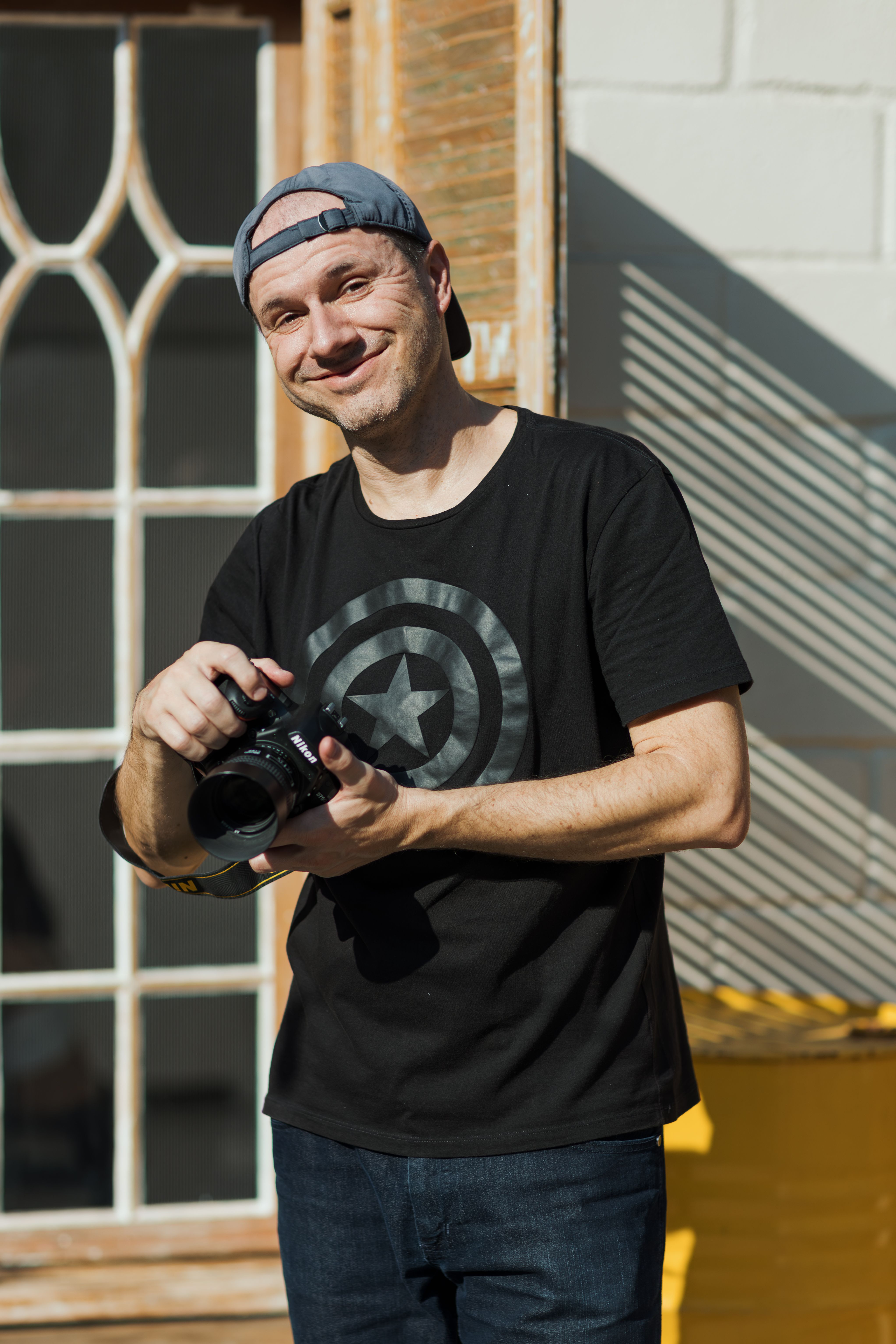Smiling man holding a camera outdoors, dressed casually with a cap.