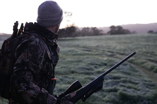 Man Wearing Gray and Black Camouflage Jacket Holding Rifle Walking on Grass Field