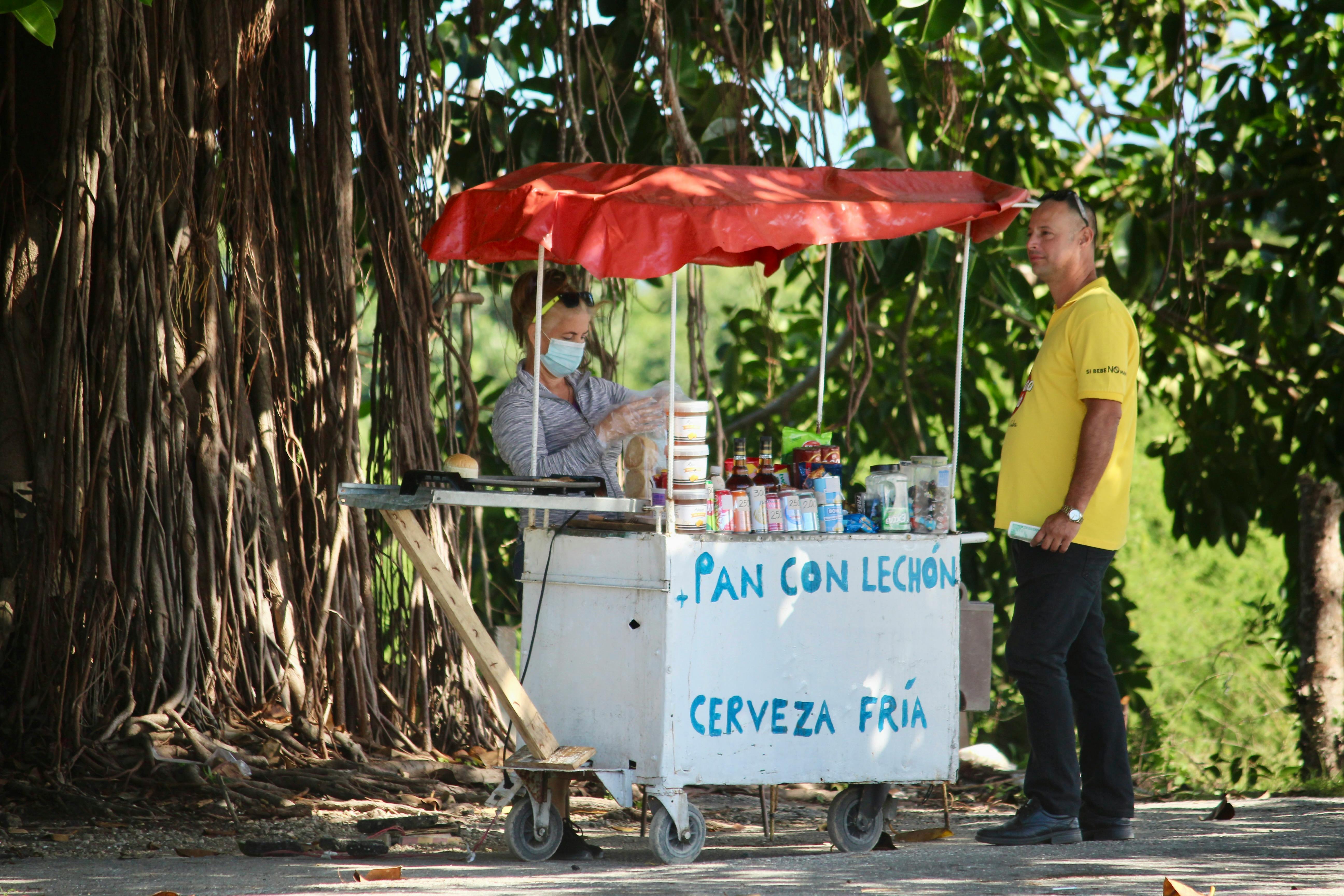 Street vendor with food cart under tree canopy offering pan con lechon and cold drinks.
