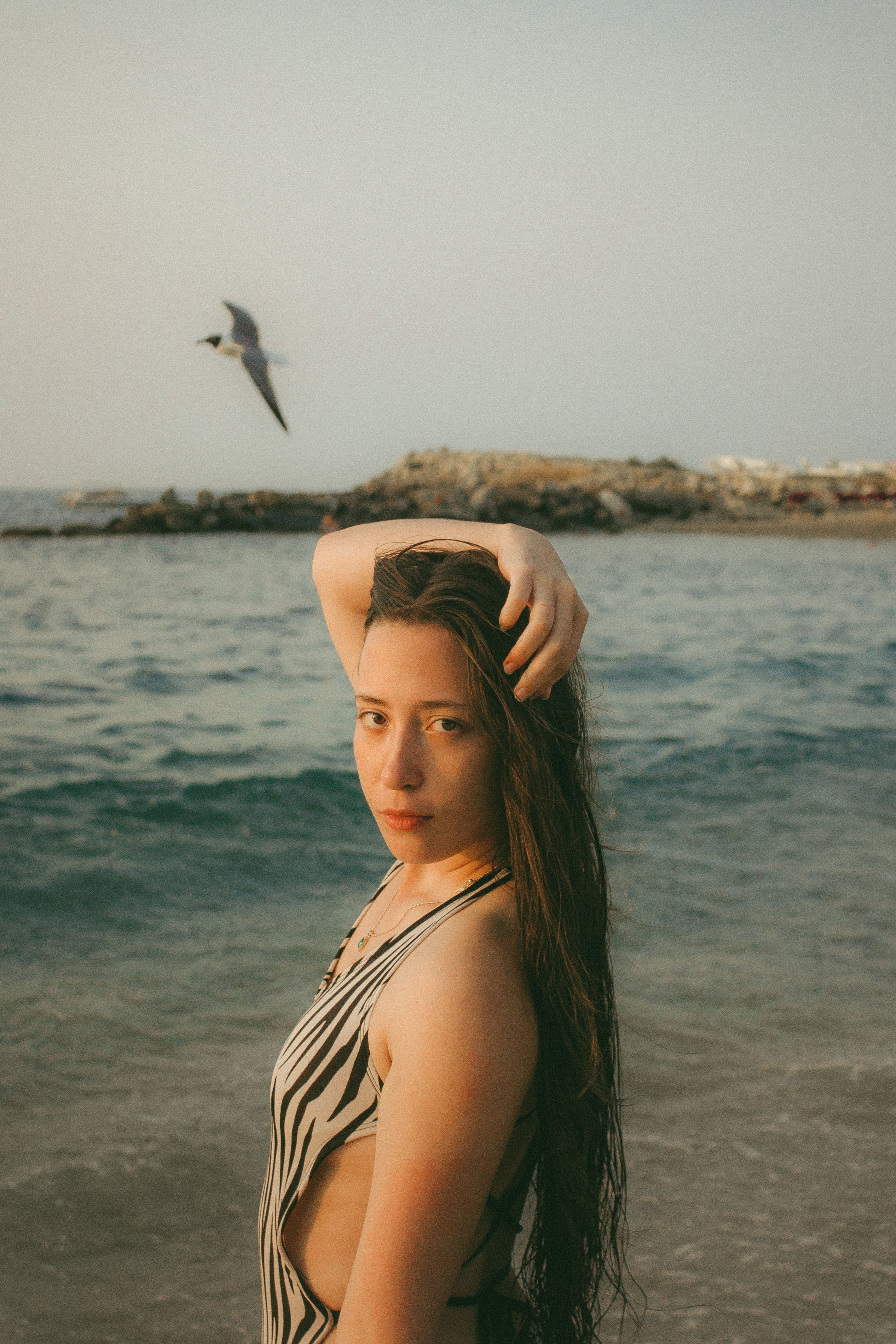 Woman in a swimsuit poses by the sea with a bird in flight, highlighting summer vibes.
