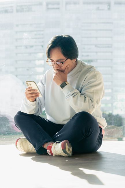 Man in casual attire sitting indoors, using smartphone against city backdrop.