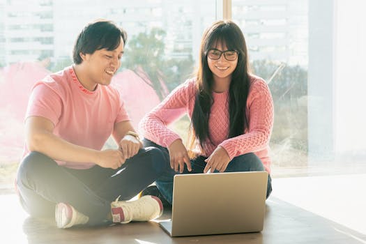 Two young adults smiling and collaborating on a laptop indoors with natural sunlight.