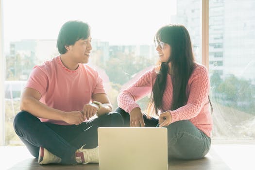 Two young adults engaged in discussion while working on a laptop near a large window.