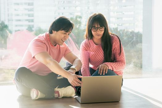 Two young adults in casual attire working together on a laptop by a window.