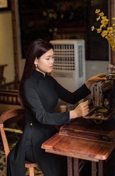 A woman in a traditional Vietnamese long dress operates a vintage sewing machine indoors.