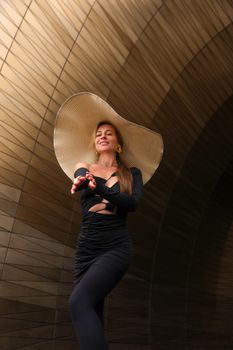 Fashionable woman poses in dramatic black dress and wide-brimmed hat against elegant architectural backdrop.