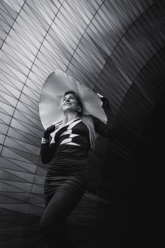 Elegant woman wearing a large hat posing confidently against modern architectural backdrop.