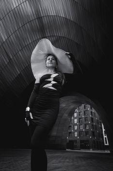 Elegant woman in dramatic hat posing under modern architectural structure.