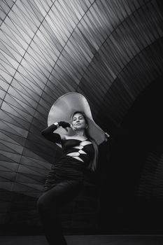 Stylish woman poses with a large hat against a sleek modern architectural background.