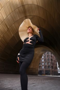 Elegant woman in black dress with a large hat poses under an architectural archway.