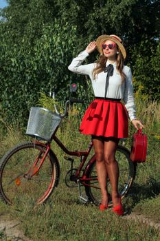 Stylish woman in red skirt posing outdoors with a bicycle and suitcase in summer.