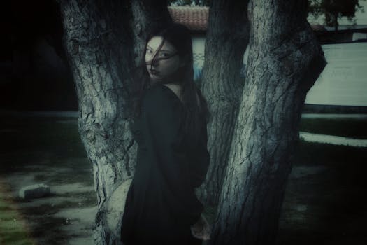 Ethereal woman in dark ambiance poses by tree, exuding gothic allure and mystery.