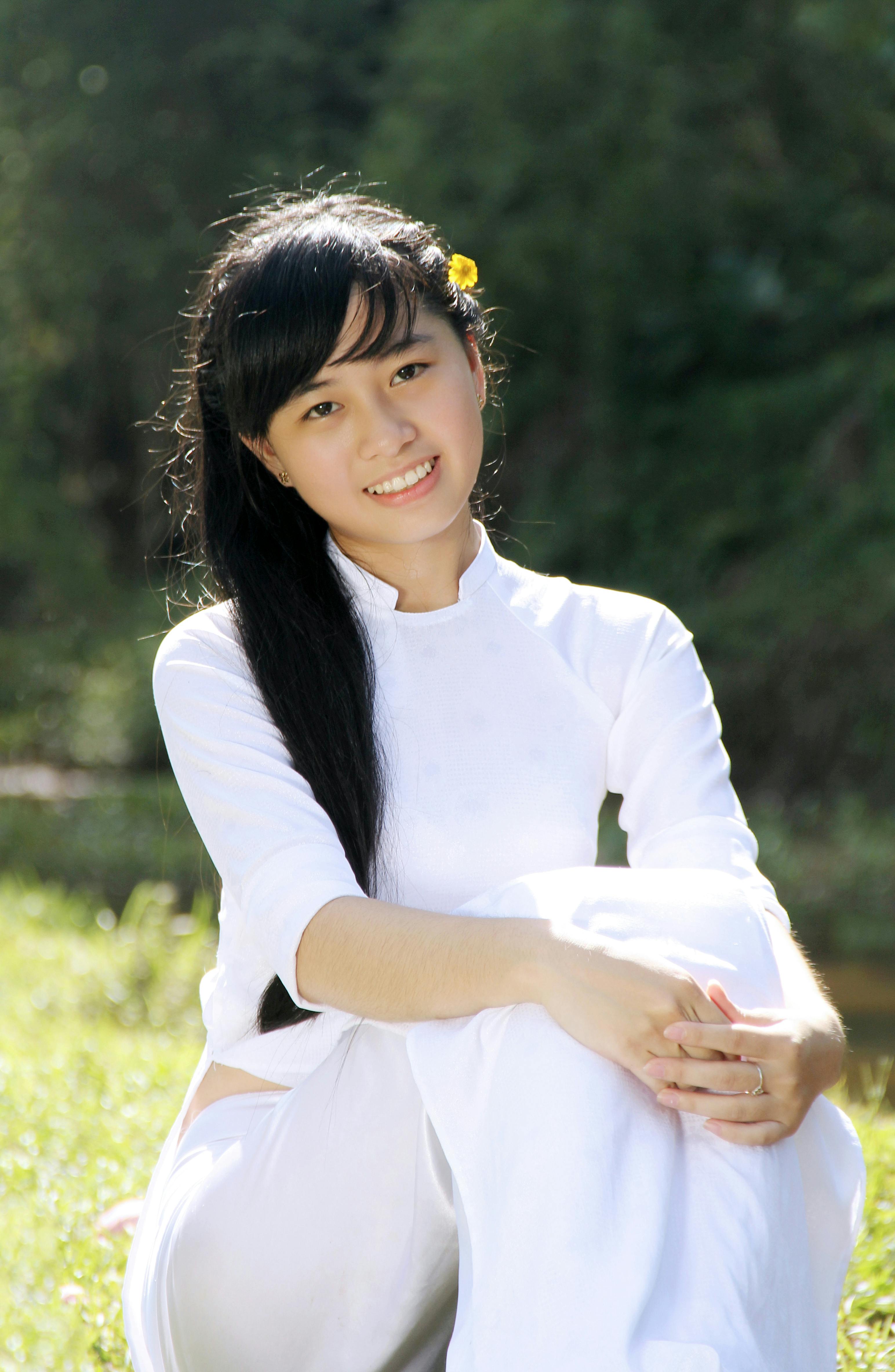 Smiling woman in white áo dài, outdoors in Hội An, Vietnam, with natural backdrop.