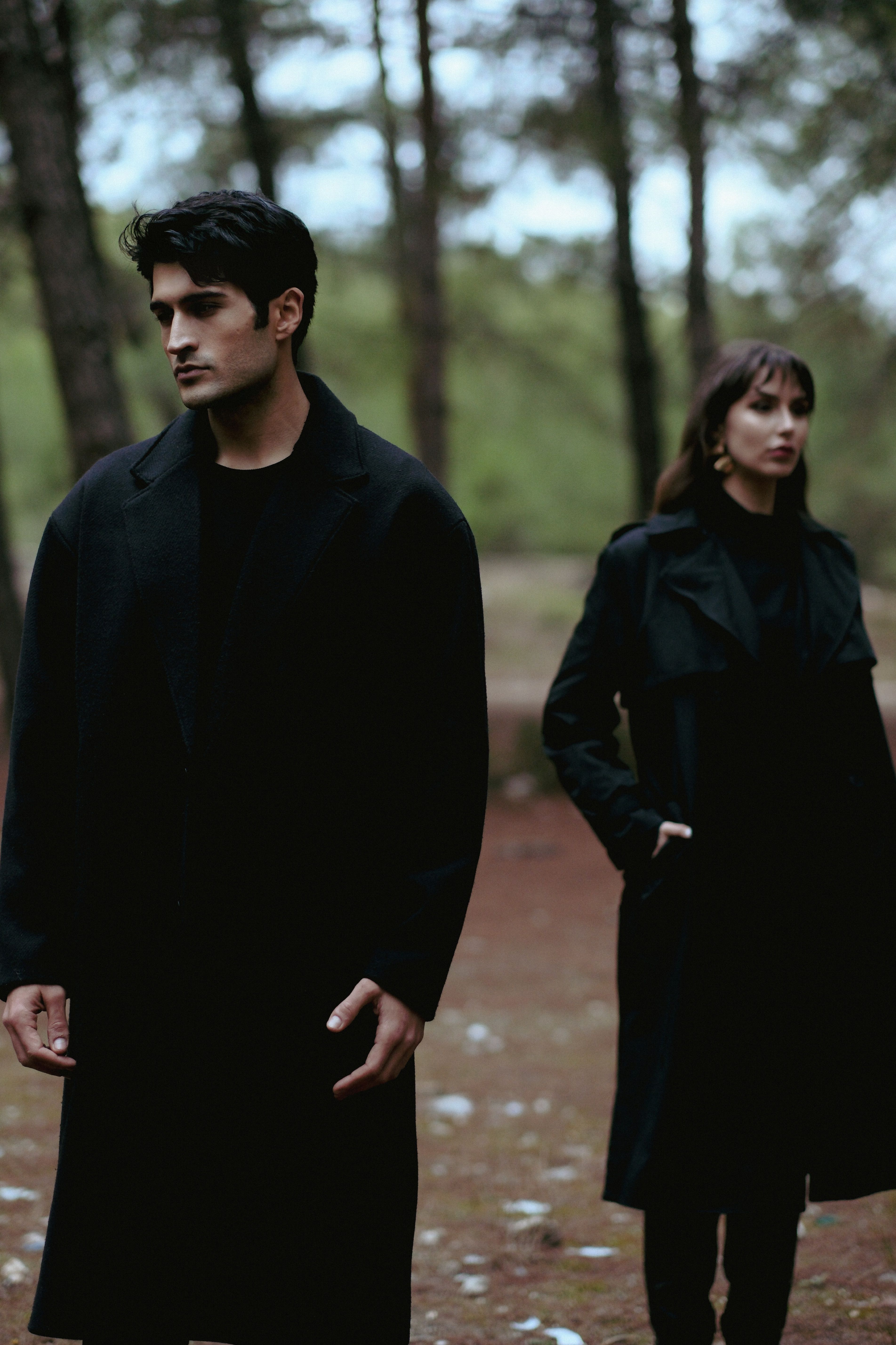 Elegant couple in long coats posing thoughtfully in a scenic forest.