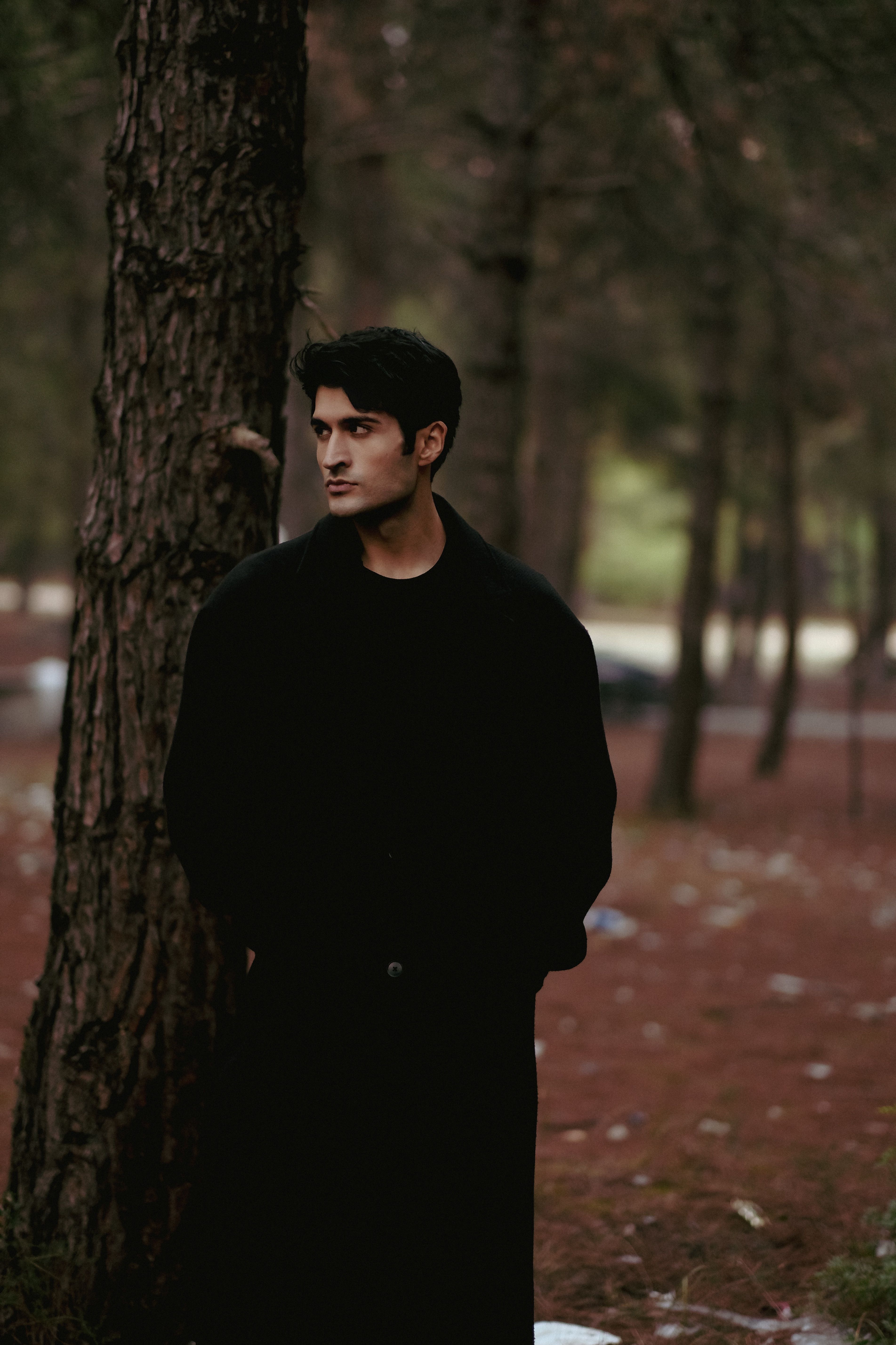 A man in a dark overcoat stands thoughtfully in a calm forest setting.
