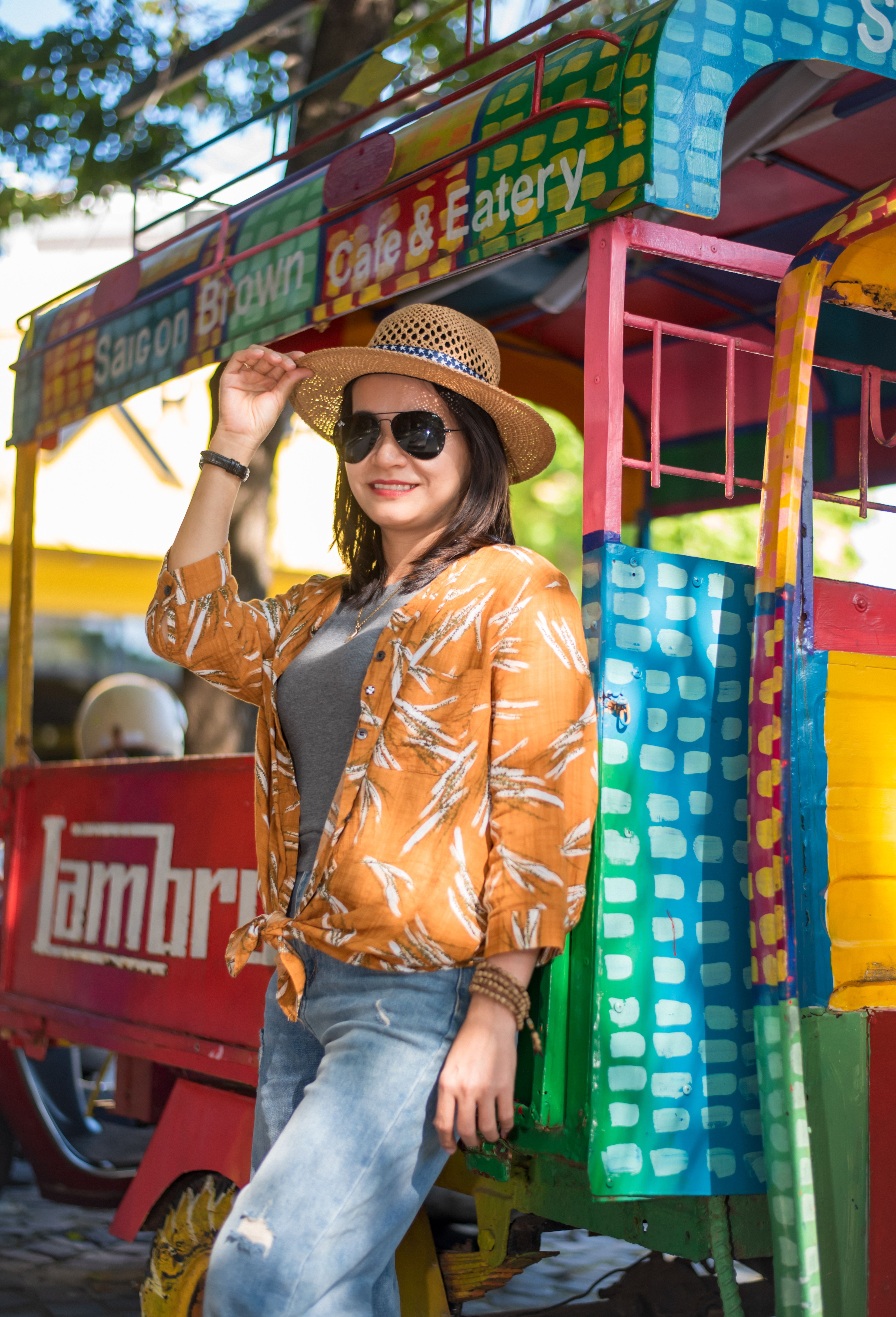 Stylish woman in sunglasses and straw hat posing by a colorful cart outdoors.