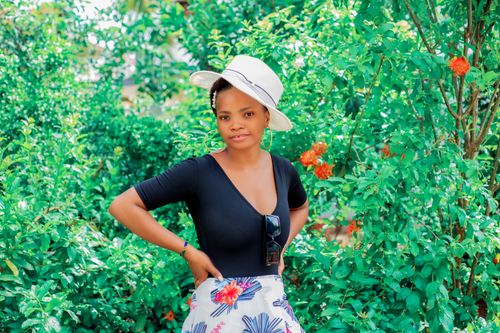 Free Confident young woman in a hat and floral skirt stands against vibrant green leaves, embracing summer vibes. Stock Photo