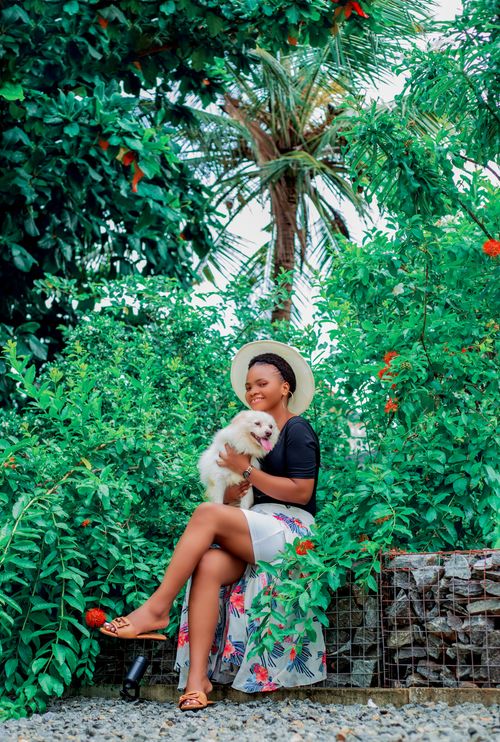 Free Smiling woman sitting amidst lush green foliage, holding her fluffy dog and wearing a sun hat. Stock Photo
