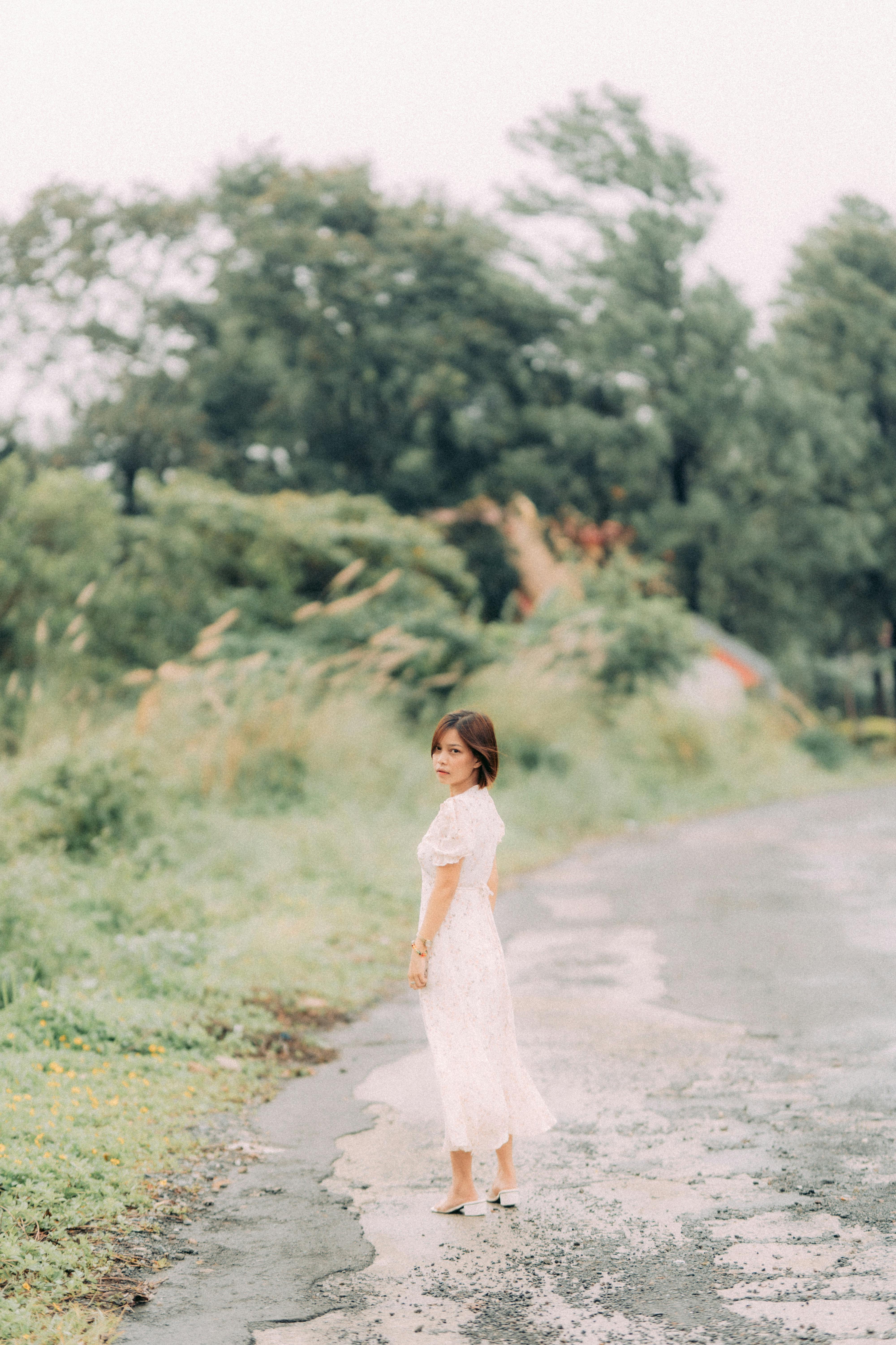A young woman in a white dress standing on a wet country road surrounded by lush greenery.