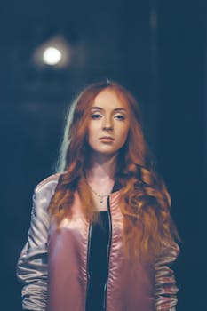 Elegant woman with red hair wearing a pink satin jacket, posing outdoors at night.