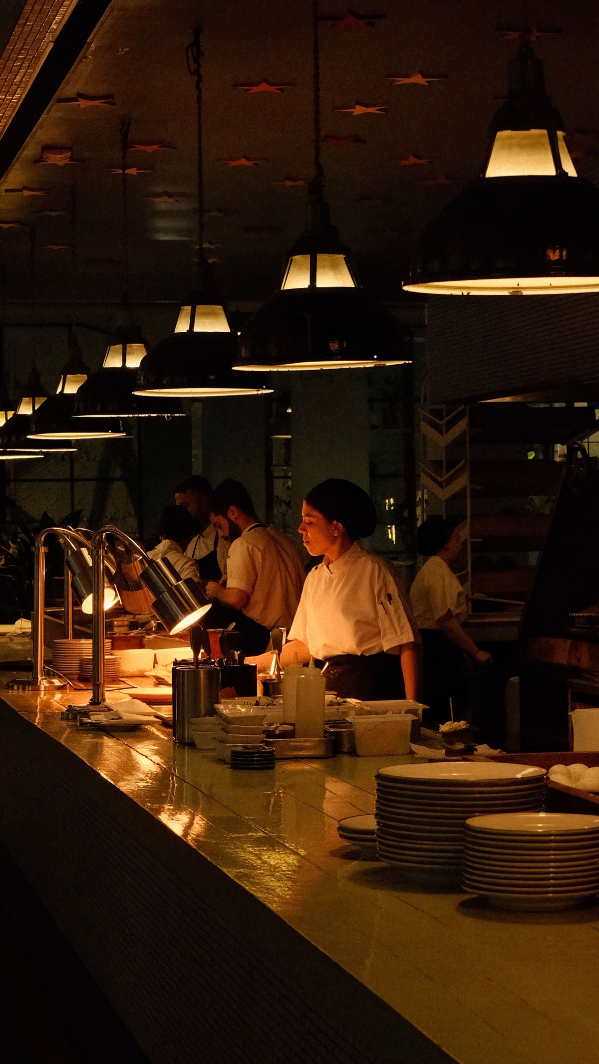 Cooks working an open kitchen under pendant lights, seen from the counter