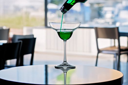 A vibrant green cocktail being poured into a glass indoors with blurred background.