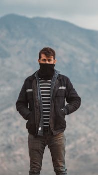 Man wearing a jacket stands outdoors with mountains in the background.