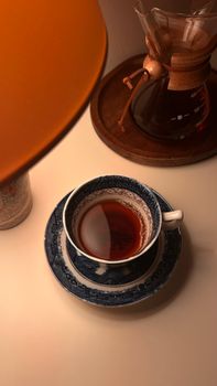 Blue and white tea cup with tea, Chemex coffee maker, and warm lamp on tabletop.