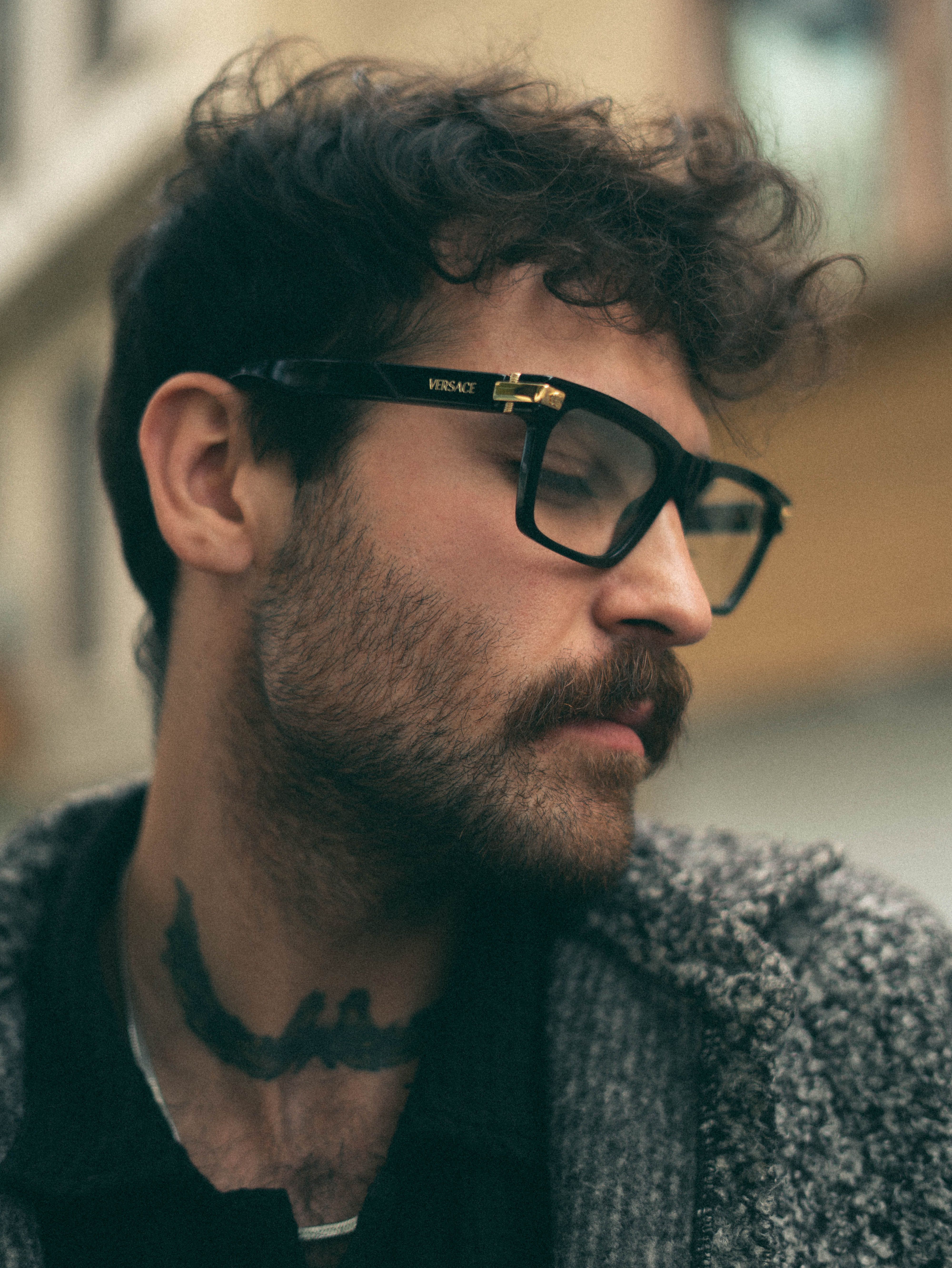 Portrait of a man with a beard and glasses, wearing a sweater, in an urban environment.