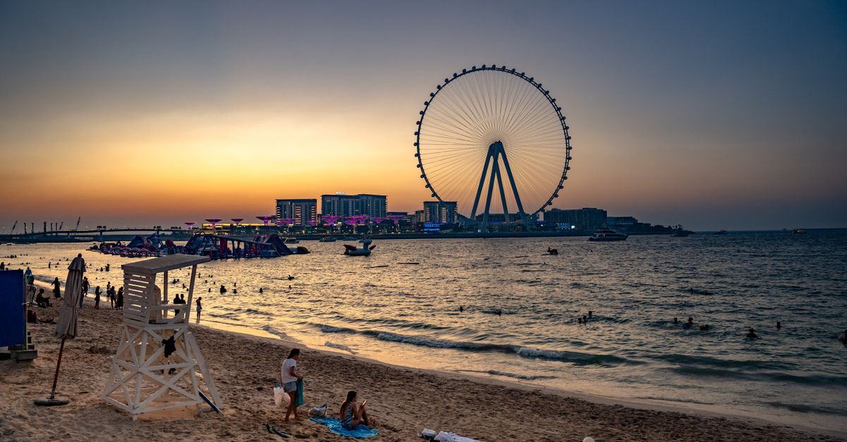 People walk through a modern Dubai shopping mall near large windows overlooking the city skyline at midday.