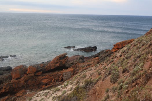 Scenic coastal cliff with red rocks and ocean waves on a cloudy day.