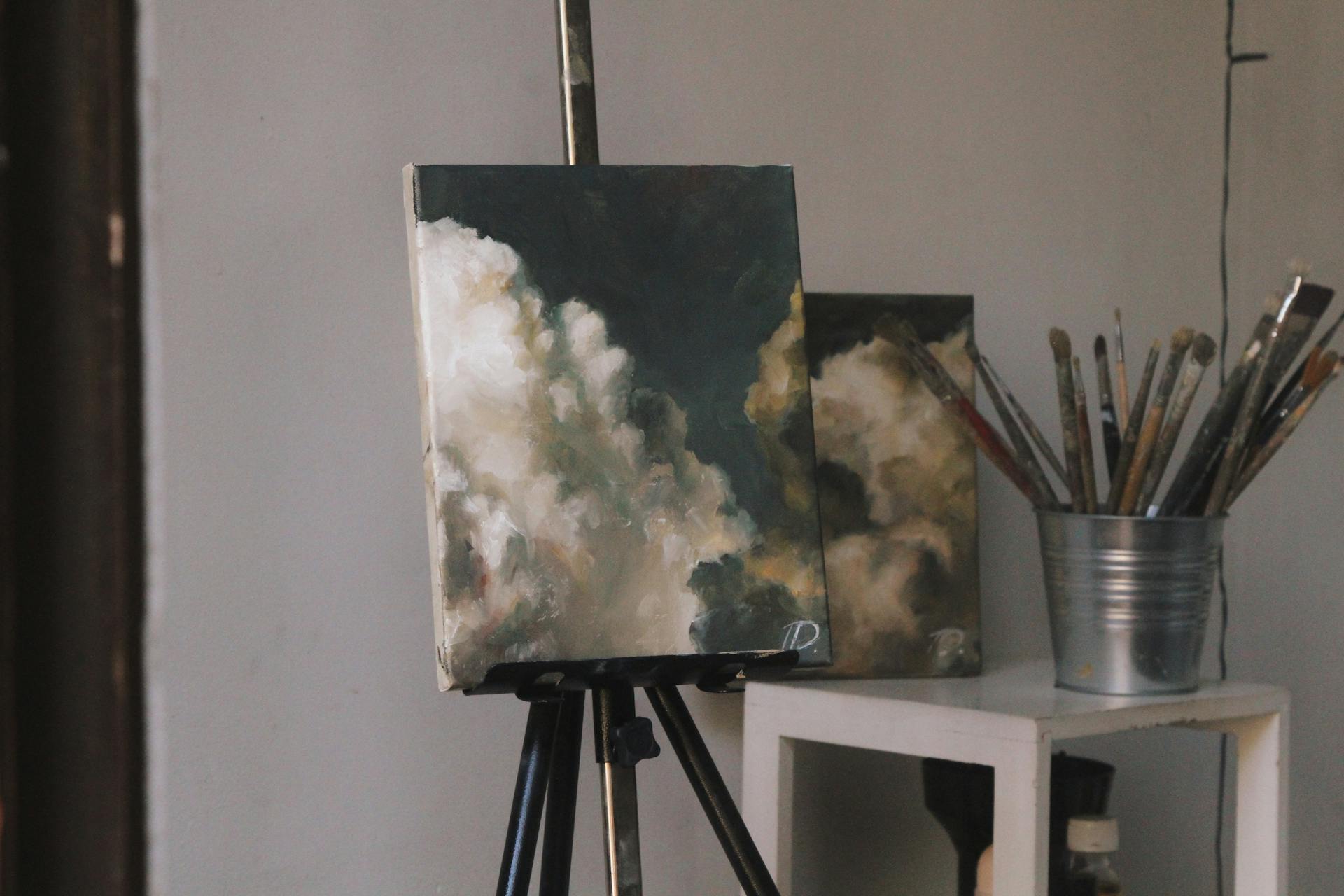 A detailed painting of clouds on an easel, placed in an art studio with brushes in a container.