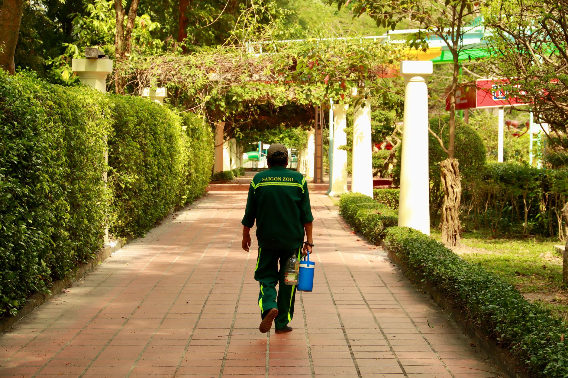 Gardener in uniform walking through Saigon Zoo's lush green pathway, carrying gardening tools.