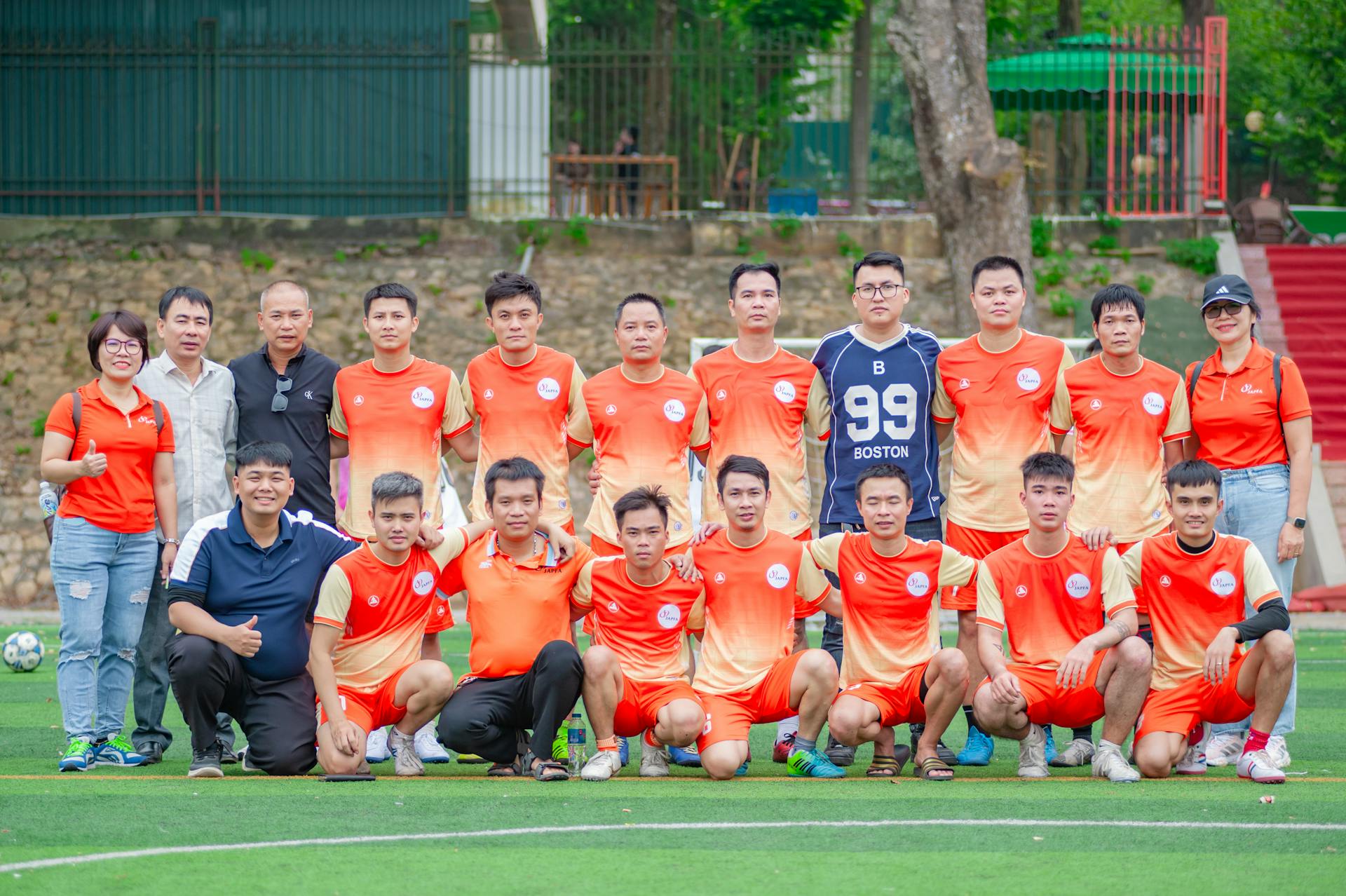 Outdoor group photo of a Vietnamese soccer team in orange uniforms at a park in Hanoi.