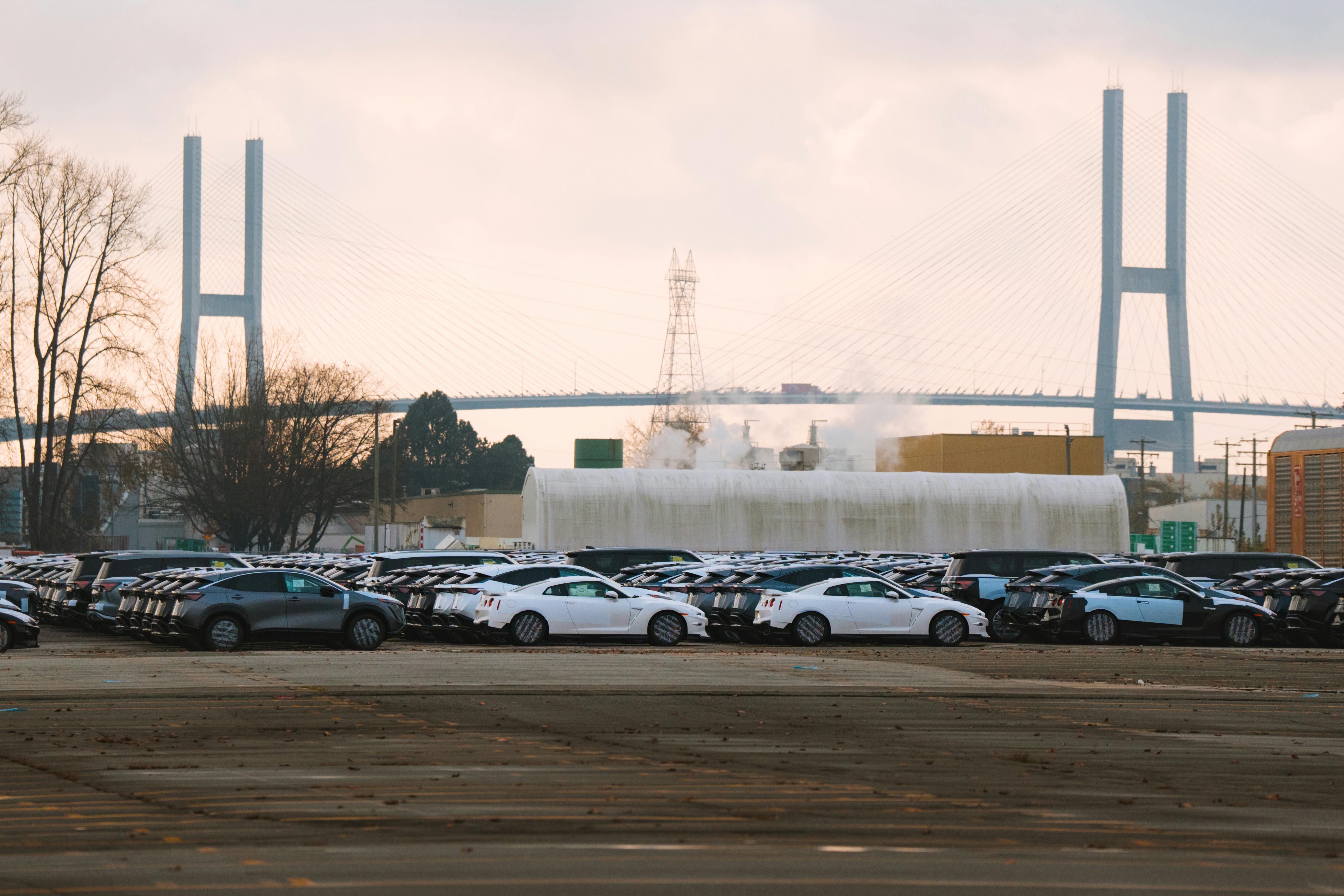 Rows of new cars at a dealership with a bridge in the background under a cloudy day.