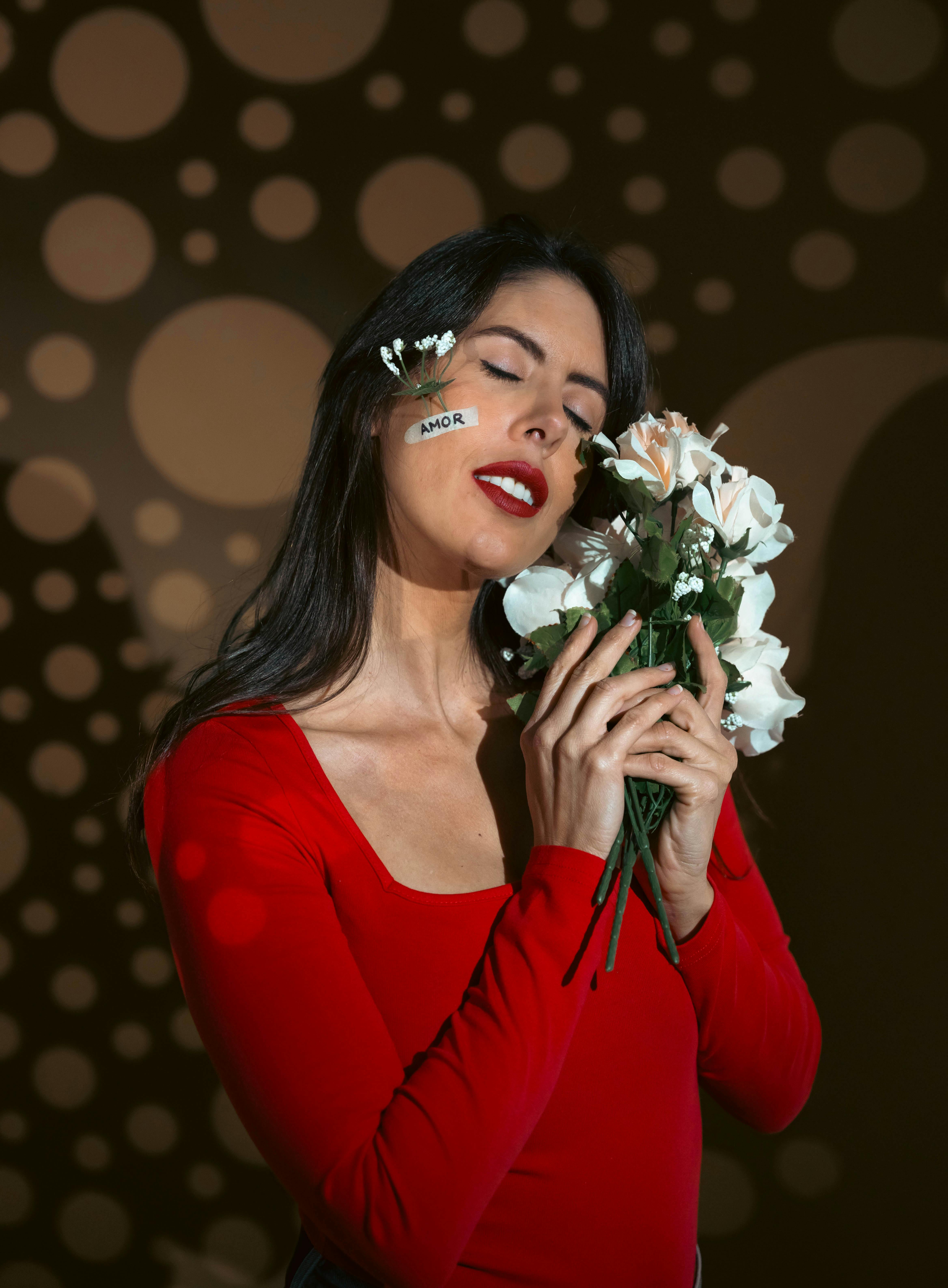 Artistic portrait of a woman in red with roses and 'Amor' text on cheek under dramatic lighting.