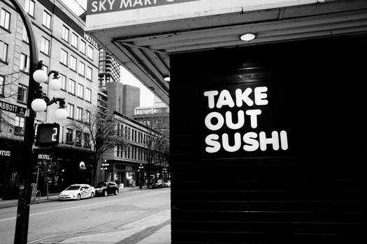 Black and white street view of a sushi takeout sign in urban setting.