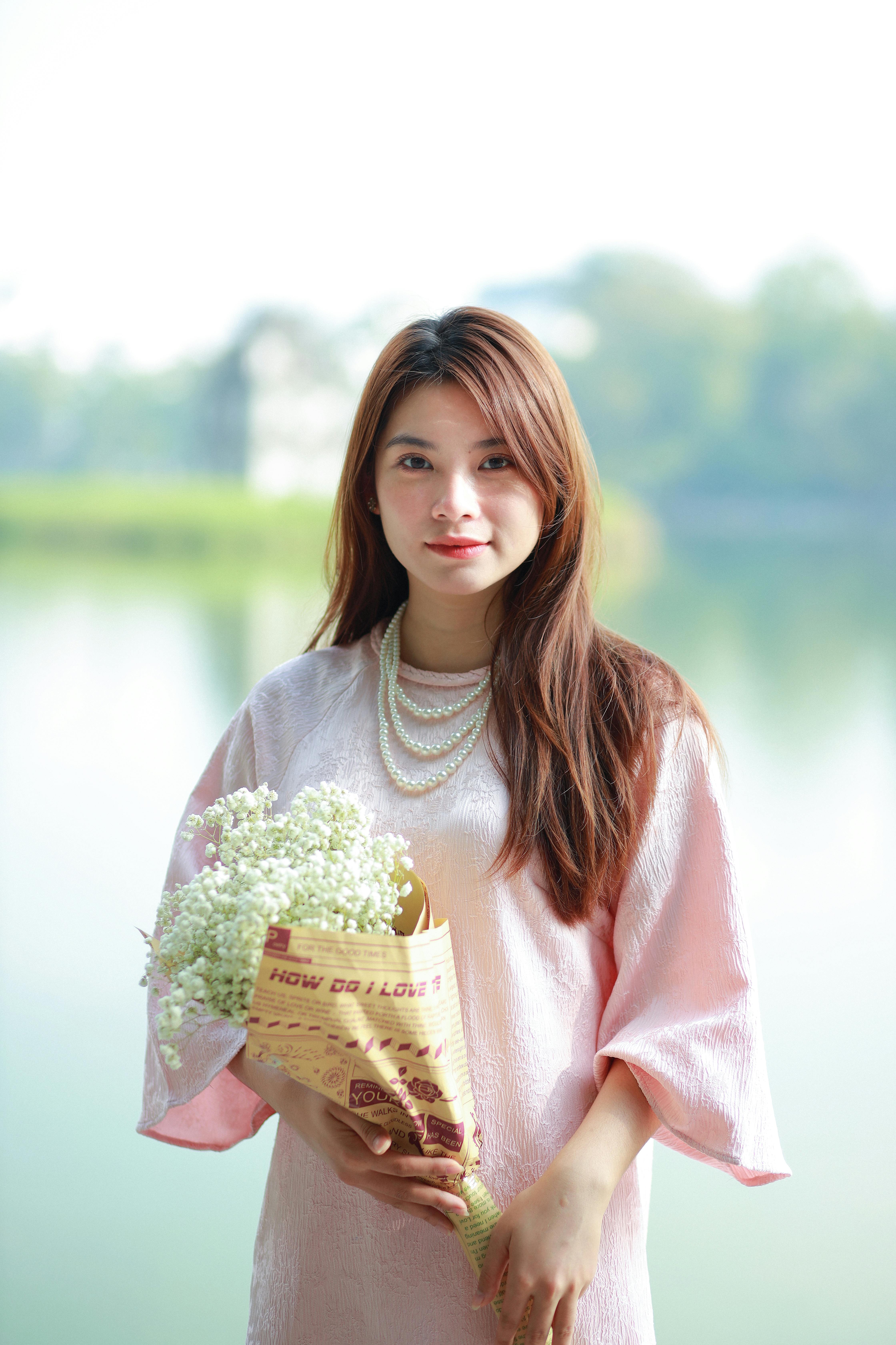 Portrait of a young woman holding flowers by a serene lake in Hanoi, Vietnam, captured on a sunny day.
