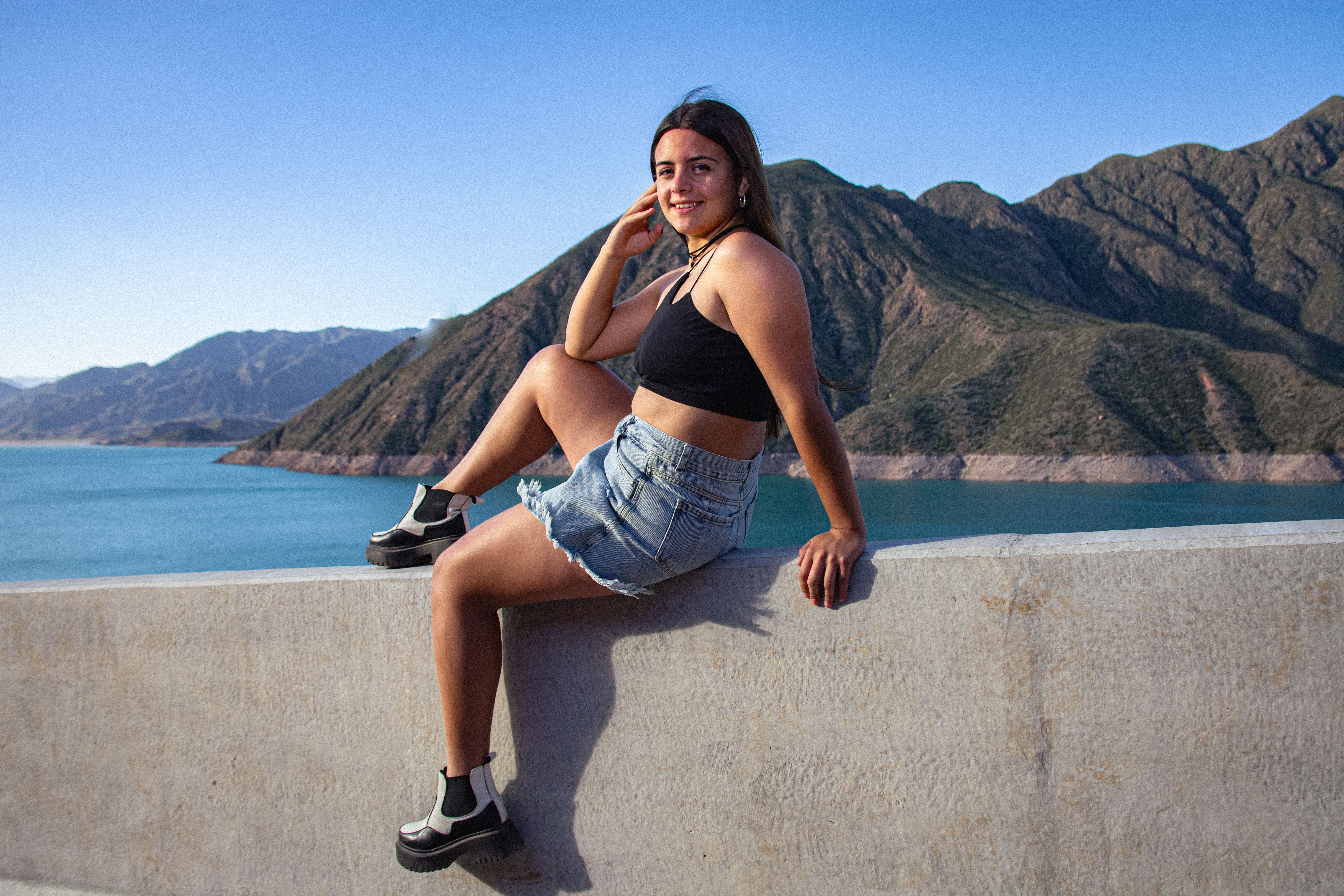 A smiling woman in casual attire sits on a ledge with a stunning mountain backdrop.