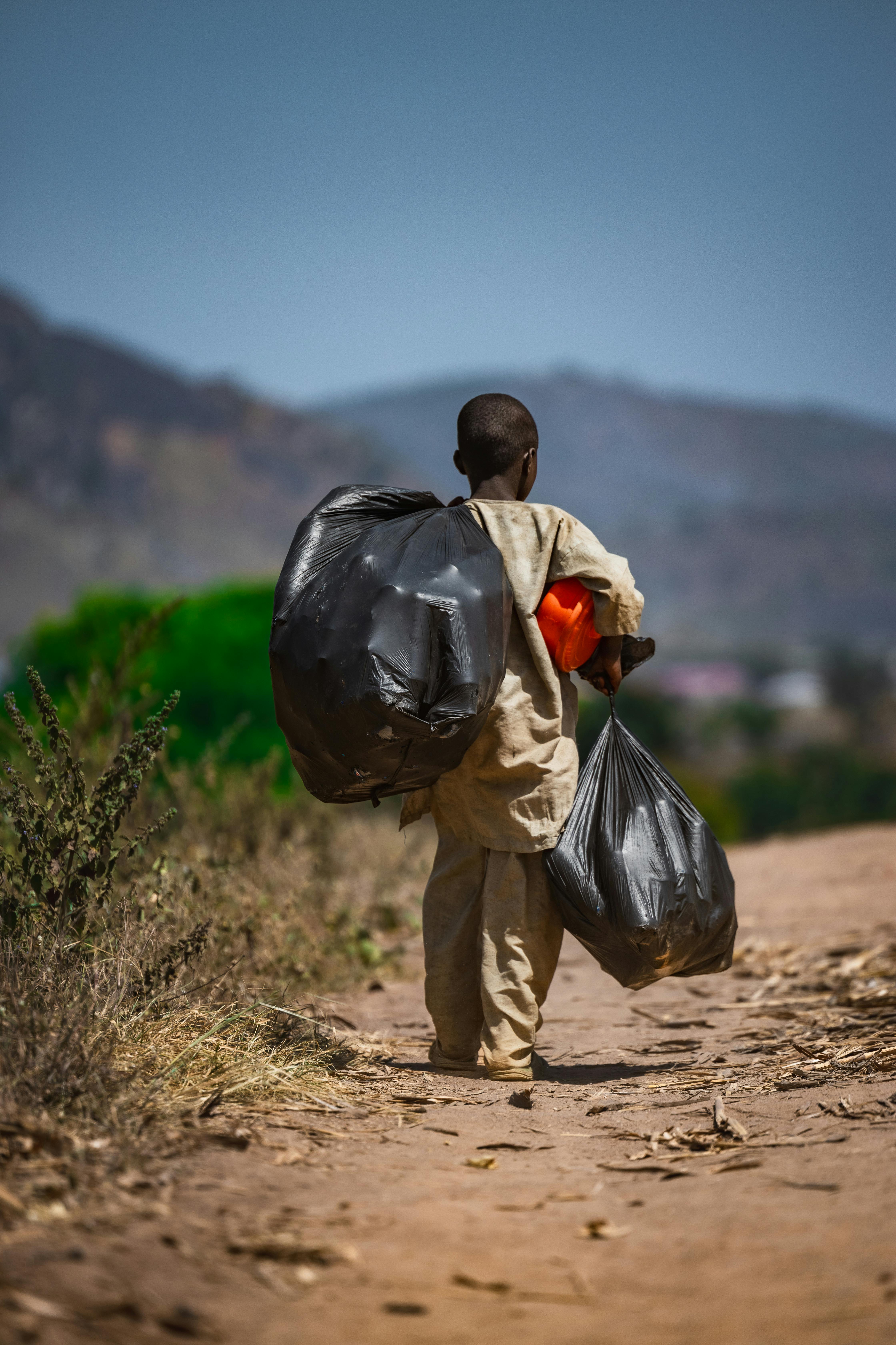 A young boy carries bags on a dirt road in Deidei, Nigeria, surrounded by mountainous terrain.